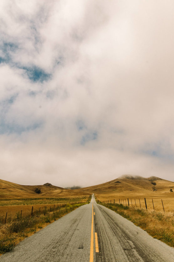 A long, empty country road stretches into the distance between golden grassy hills under a partly cloudy sky. Fence lines run parallel to the road on both sides. by Justin Salem Meyer