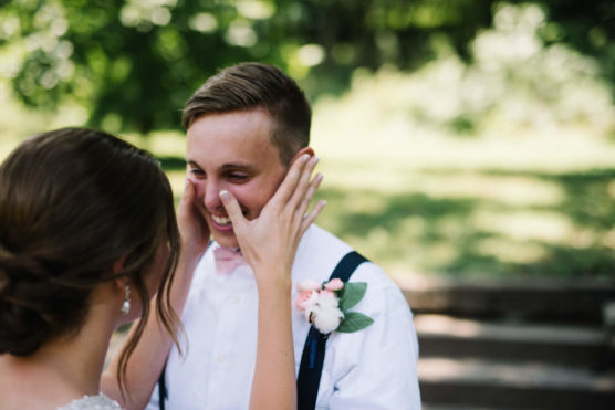 A person in a white shirt and suspenders smiles joyfully as another person gently holds their face with both hands; the background is bright and green, suggesting an outdoor setting. by Justin Salem Meyer