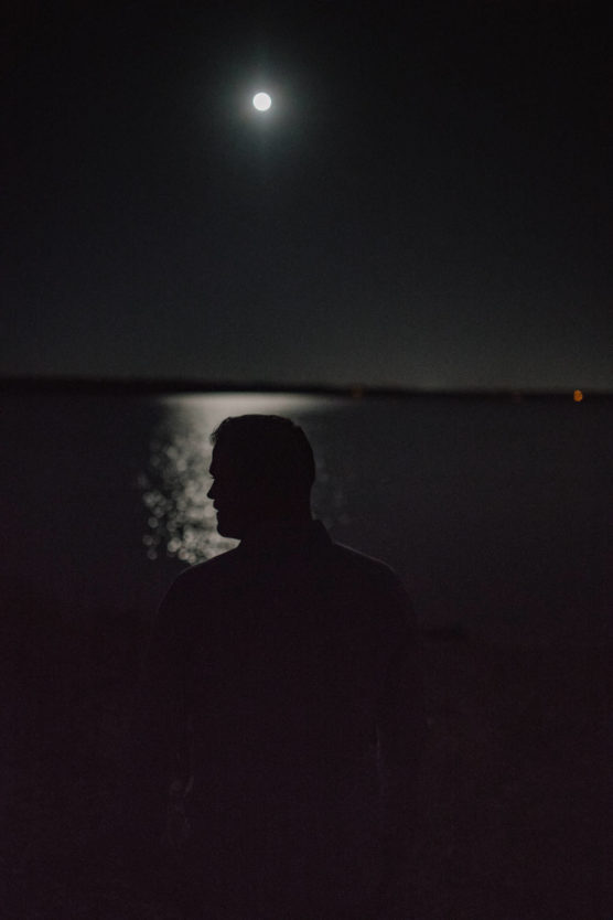 A silhouette of a person stands by a body of water at night, with the full moon shining brightly above and its reflection shimmering on the water’s surface. by Justin Salem Meyer