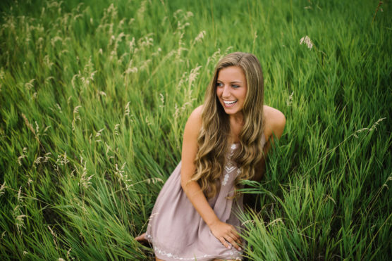 A young woman with long, wavy hair wearing a light pink dress is sitting and smiling in a field of tall green grass. by Justin Salem Meyer