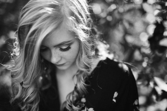 Black and white photo of a young woman with long, wavy hair looking down, surrounded by blurred foliage. She wears a dark, patterned top and her face is softly lit with delicate shadows. by Justin Salem Meyer