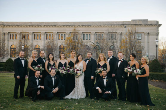 A wedding party poses outdoors in front of a large, historic building. The group includes the bride in a white gown, bridesmaids in black dresses, and groomsmen in black suits with bow ties. Everyone is smiling and holding flowers. by Justin Salem Meyer
