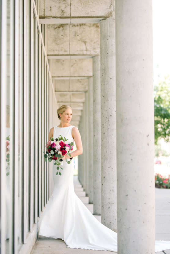 A bride in a white sleeveless gown stands by tall concrete columns, holding a bouquet of red, pink, and purple flowers with greenery, looking to her left in natural daylight. by Justin Salem Meyer