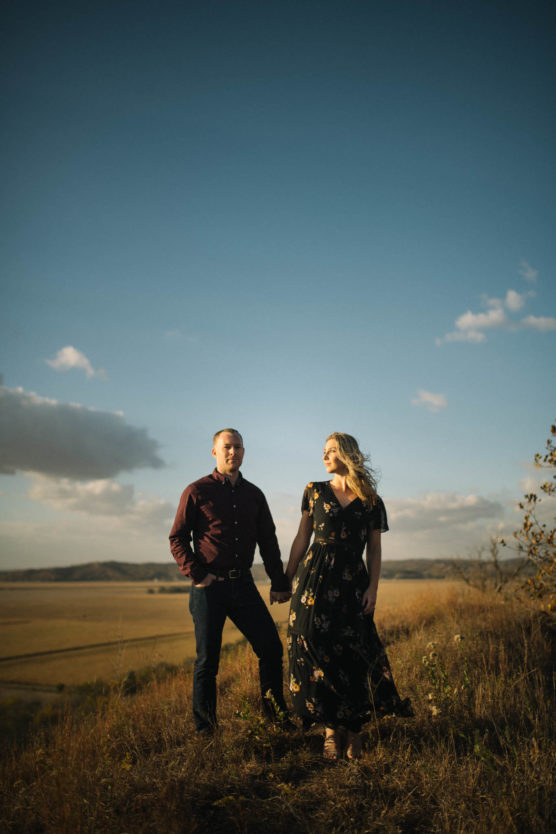 A man and a woman stand holding hands on a grassy hill at sunset, with open fields and distant hills in the background. The man wears a maroon shirt and dark pants; the woman wears a black floral dress. by Justin Salem Meyer