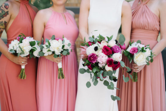 Four women in pink and cream dresses hold bouquets of white, pink, and purple flowers with greenery. The woman in white holds a larger bouquet; their faces are not visible. by Justin Salem Meyer