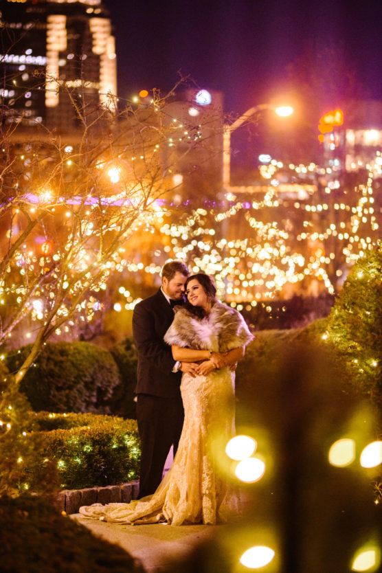 A couple dressed in formal attire embraces outdoors at night, surrounded by festive string lights and glowing city buildings in the background. The woman wears a gold dress and fur shawl; the man is in a black suit. by Justin Salem Meyer