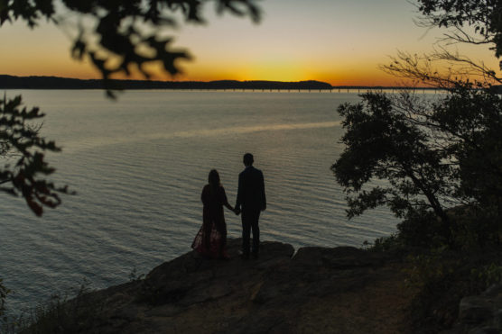 A couple stands hand in hand on a rocky cliff, overlooking a calm lake at sunset, with a distant bridge visible and trees framing the scene. by Justin Salem Meyer