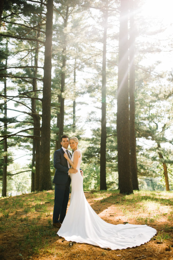A bride in a white gown and a groom in a dark suit stand close together in a sunlit forest, surrounded by tall pine trees with sunlight filtering through the branches. by Justin Salem Meyer