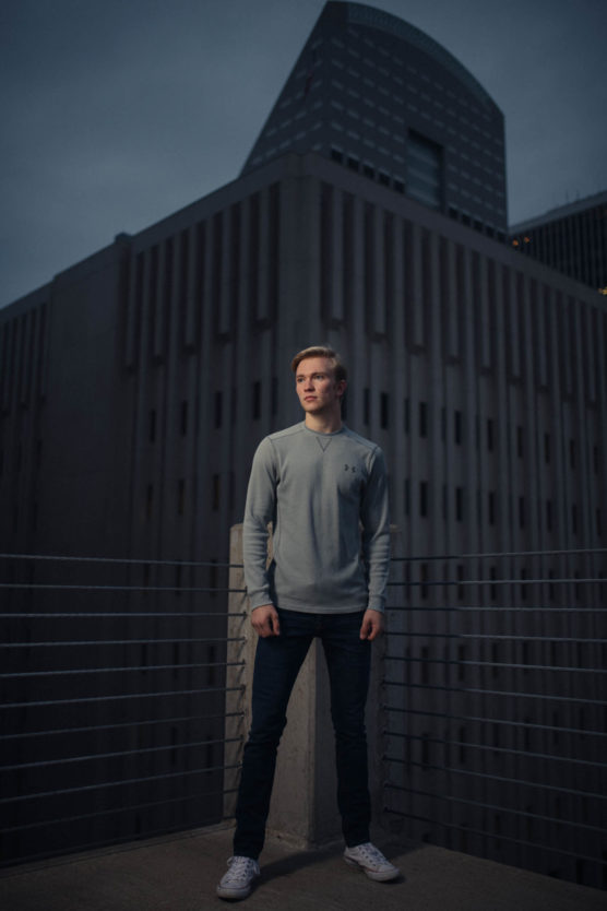 A young man stands on a rooftop at dusk, wearing a light gray sweater, dark jeans, and white sneakers. Tall, modern buildings with geometric patterns rise behind him in the dim, moody lighting. by Justin Salem Meyer