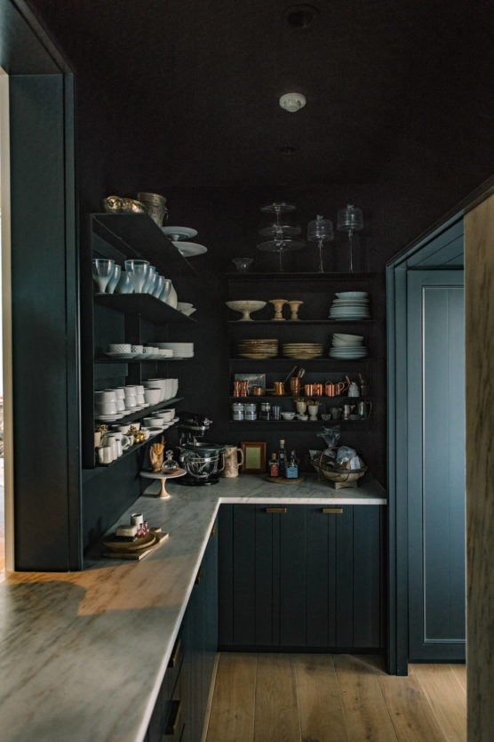 A modern pantry with dark cabinets and open shelves displaying neatly stacked plates, bowls, glassware, and kitchen appliances. A white marble countertop runs along the left side, with wooden flooring below. by Justin Salem Meyer