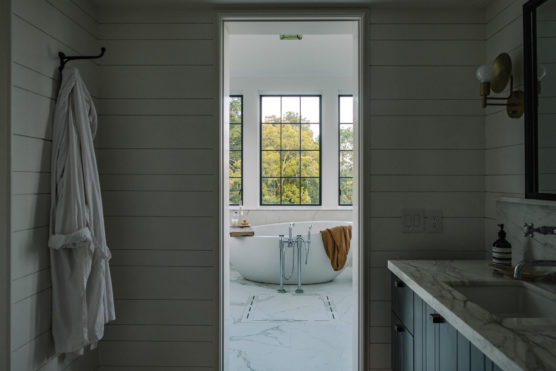 A modern bathroom with white shiplap walls, a marble countertop, and a robe hanging on a hook. Through an open doorway, a freestanding bathtub sits by large windows overlooking trees. by Justin Salem Meyer