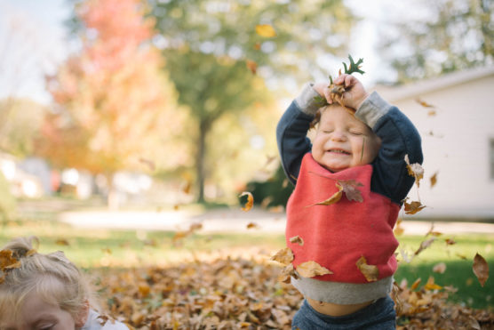 A young child wearing a red sweater smiles joyfully with eyes closed, arms raised, and autumn leaves falling around them outdoors. The background shows trees with colorful foliage and a house. by Justin Salem Meyer