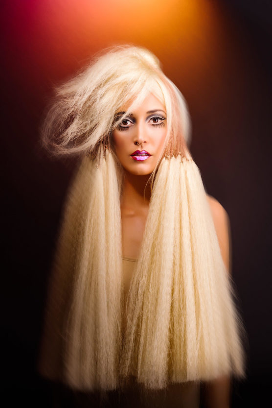 A woman with dramatic makeup and long, voluminous, platinum blonde hair styled in textured, crimped waves, stands against a dark gradient background with orange and pink light above her head. by Justin Salem Meyer