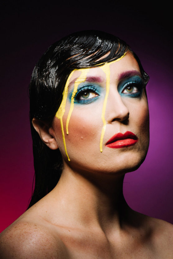 A person with slicked-back dark hair, bold blue and pink eye makeup, red lipstick, and vibrant yellow paint streaks running down their face, posing against a purple and pink gradient background. by Justin Salem Meyer