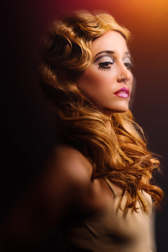 A woman with long, wavy blonde hair and dramatic makeup, including bold eyeliner and pink lipstick, poses in profile against a dark background with a warm light source in the upper right corner. by Justin Salem Meyer