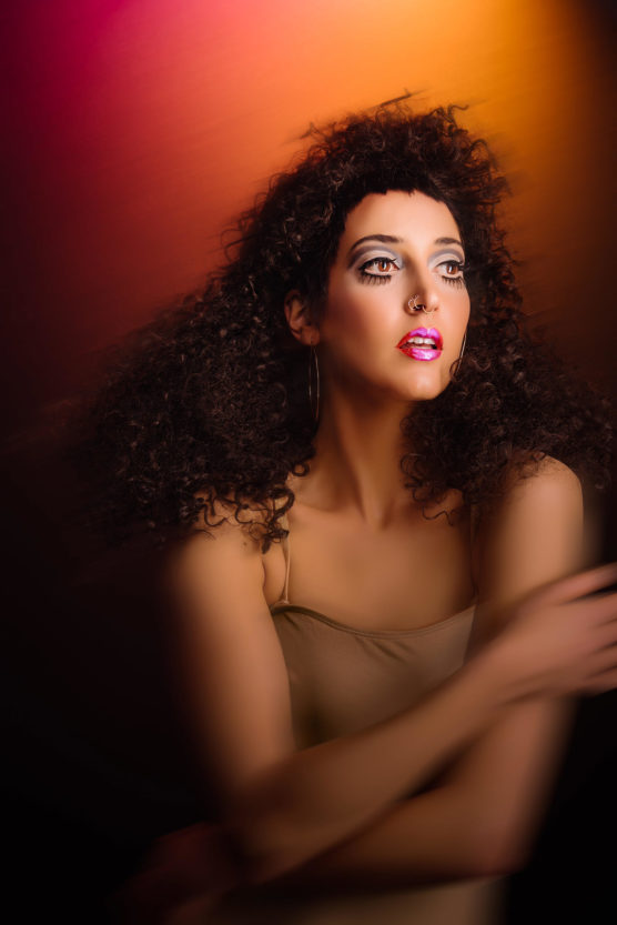 A person with curly dark hair, dramatic eye makeup, and glossy pink lips poses against a vibrant, colorful background. They wear a beige top and look upwards with one arm crossed in front. The lighting is soft and artistic. by Justin Salem Meyer