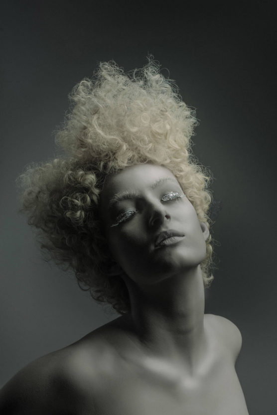 A person with pale skin and dramatic, curly platinum blonde hair poses with closed eyes and white eyelash makeup, set against a dark, neutral background. Their expression is serene and artistic. by Justin Salem Meyer