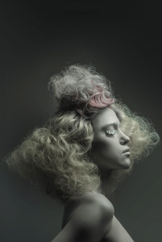 A person with voluminous, curly platinum blonde hair styled in an avant-garde fashion poses in profile against a dark background. Their skin is pale, with dramatic eye makeup featuring crystal embellishments. by Justin Salem Meyer
