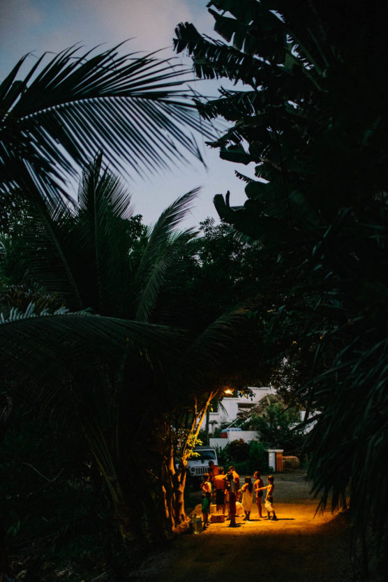 A group of people stands under a streetlight on a path surrounded by large tropical plants at dusk, with soft yellow light illuminating them against the dark, leafy background. by Justin Salem Meyer
