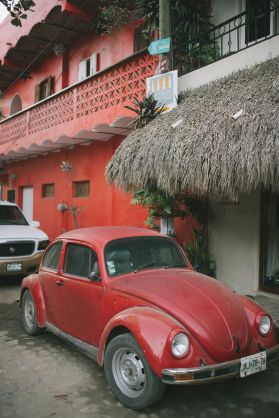 A classic red Volkswagen Beetle parked on a street in front of a vibrant red building with a thatched roof and decorative plants. A white vehicle is partially visible behind the Beetle. by Justin Salem Meyer