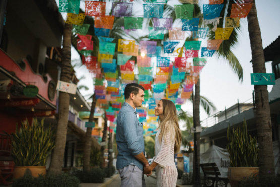 A couple holding hands and smiling at each other stands under colorful papel picado banners on a lively street lined with palm trees and shops. by Justin Salem Meyer