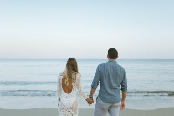 A couple holding hands stands at the edge of a calm beach, facing the ocean. The woman wears a white dress with an open back, and the man wears a blue shirt and light pants. The sky above is clear and serene. by Justin Salem Meyer