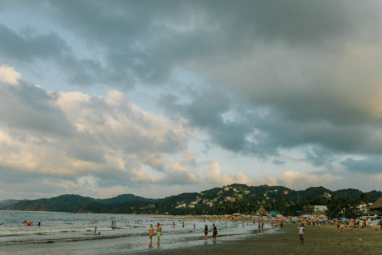 A wide sandy beach with many people swimming, walking, and relaxing near the water. Green hills and houses are visible in the background under a sky filled with clouds. by Justin Salem Meyer