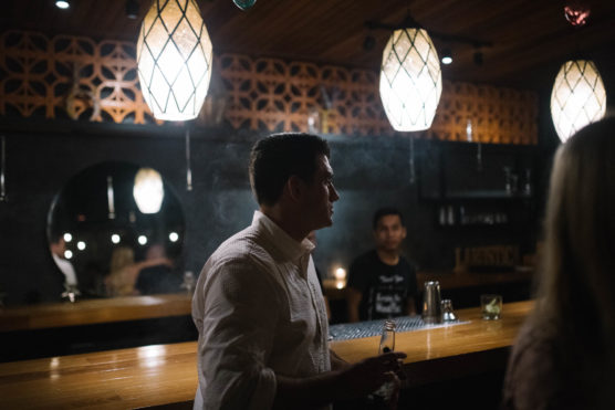 A man in a white shirt stands at a dimly lit bar with warm hanging lights. Another person sits behind the bar, and a few drinks are on the counter. The atmosphere feels relaxed and intimate. by Justin Salem Meyer