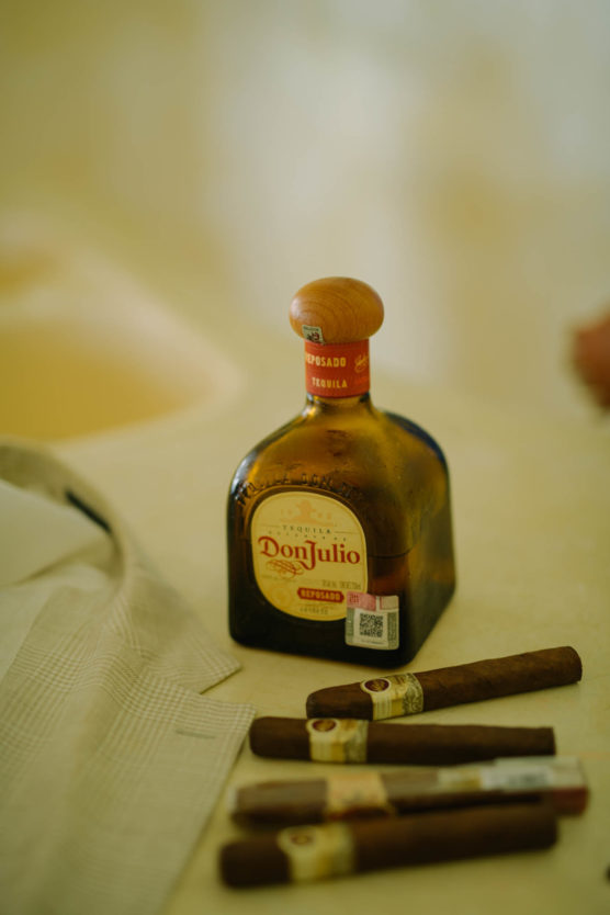 A bottle of Don Julio tequila sits on a table next to three cigars and a folded light-colored cloth or jacket. The background is softly blurred. by Justin Salem Meyer