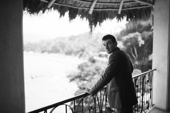 A man in a suit stands on a balcony with a thatched roof, looking back toward the camera. Trees and a blurred landscape are visible in the background. The image is in black and white. by Justin Salem Meyer