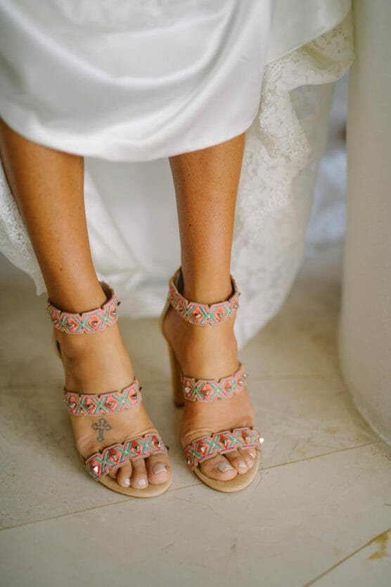 A person wearing a white dress shows off their feet in tan block-heeled sandals with colorful, embroidered straps, and a small cross tattoo on one foot. The background includes a light stone floor and lace fabric. by Justin Salem Meyer