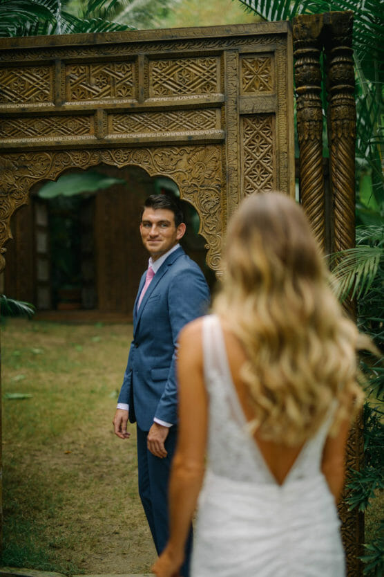 A man in a blue suit and pink tie stands in front of an ornate wooden archway, looking back at a woman in a white dress with wavy blonde hair, outdoors among lush greenery. by Justin Salem Meyer