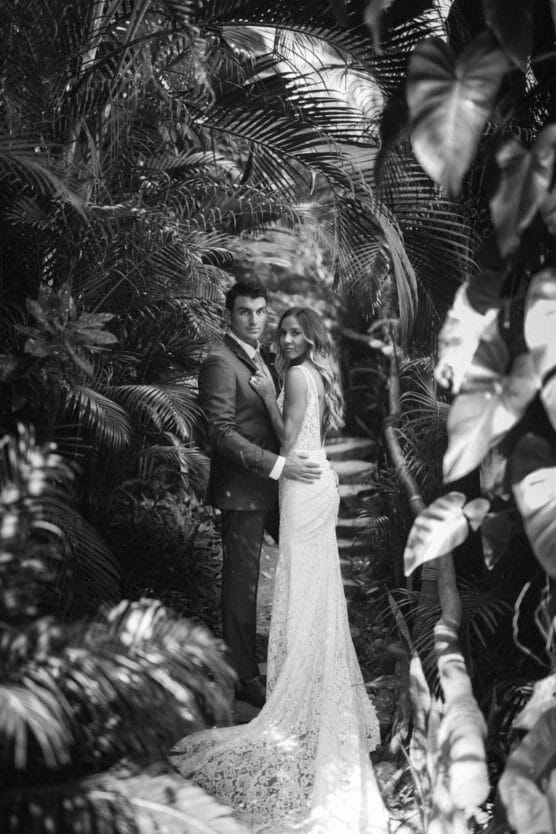 A bride and groom stand closely together on a stone path surrounded by lush tropical foliage, looking back toward the camera. The bride wears a lace gown, and the photo is in black and white. by Justin Salem Meyer