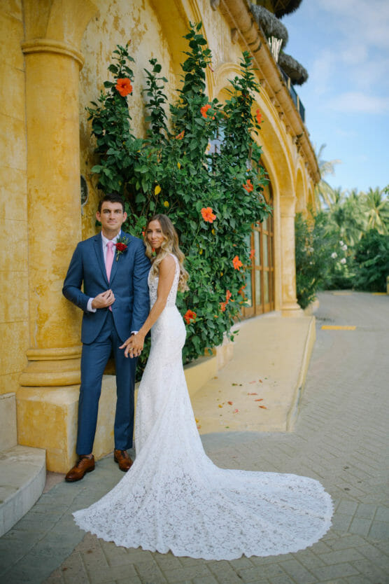 A bride in a white lace gown and a groom in a blue suit stand hand in hand by a yellow stucco building with arches and flowering vines, posing for a wedding photo outdoors. by Justin Salem Meyer