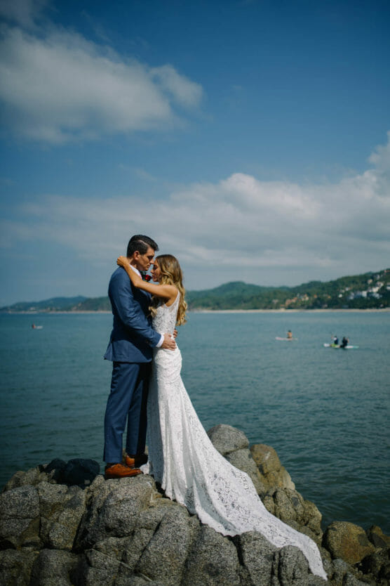 A bride and groom embrace on rocky shore, with the bride in a white lace dress and the groom in a blue suit. The ocean and distant hills are in the background under a blue sky with clouds. by Justin Salem Meyer