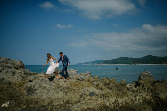 A bride in a white dress and a groom in a blue suit walk hand-in-hand over rocky terrain by the ocean, with distant boats and green hills under a blue sky. by Justin Salem Meyer