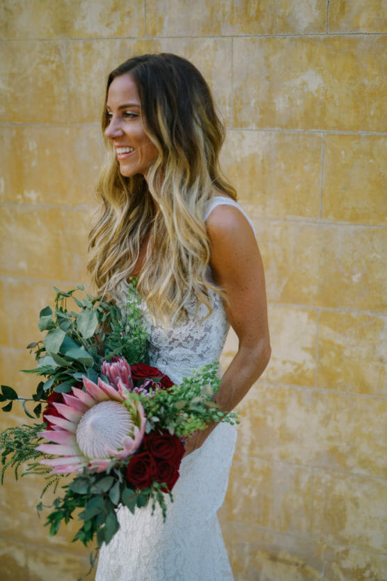 A smiling bride in a lace wedding dress holds a large bouquet with red roses, greenery, and a pink king protea, standing against a textured yellow wall. by Justin Salem Meyer
