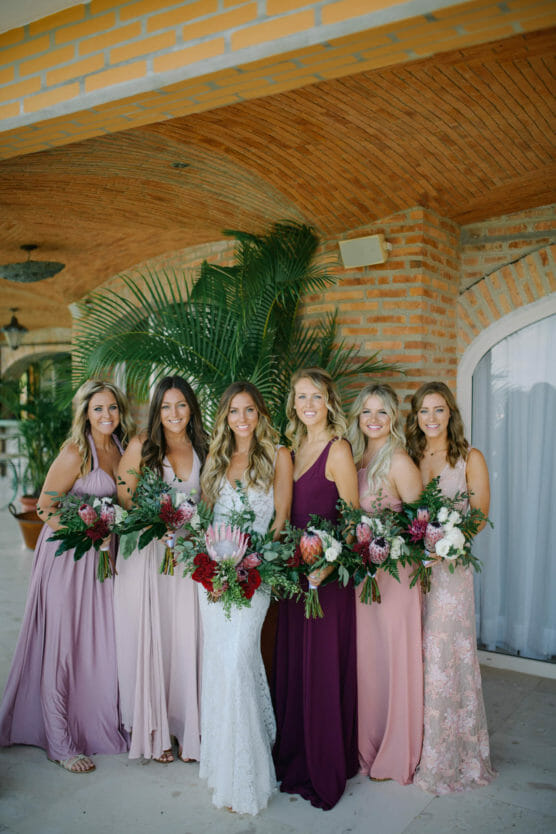 Six women in long, elegant dresses stand together, holding large floral bouquets. One woman in the center wears a white gown; the others wear pink and purple dresses. They pose and smile in front of a brick wall with greenery. by Justin Salem Meyer