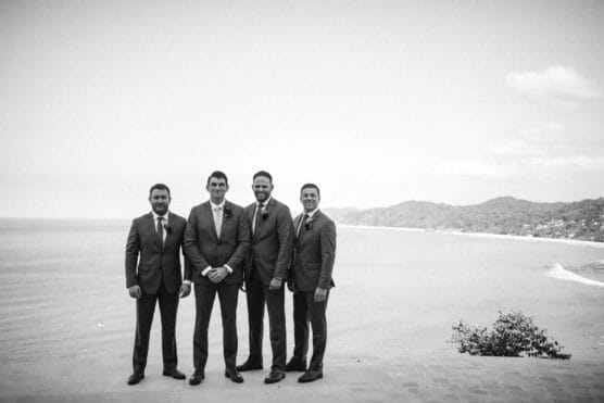 Four men in suits stand side by side on a stone terrace overlooking the ocean, with hills and coastline visible in the background. The photo is black and white. by Justin Salem Meyer
