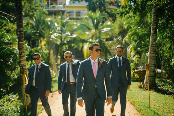 Four men in suits, wearing sunglasses, walk confidently down a sunlit path surrounded by lush greenery and palm trees, with one man leading in front. by Justin Salem Meyer