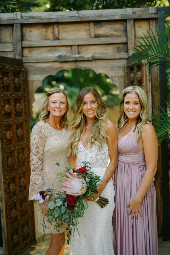Three women stand together, smiling in front of a rustic wooden arch. The central woman in a white lace dress holds a bouquet; the women on either side wear light-colored dresses. Lush greenery surrounds them. by Justin Salem Meyer