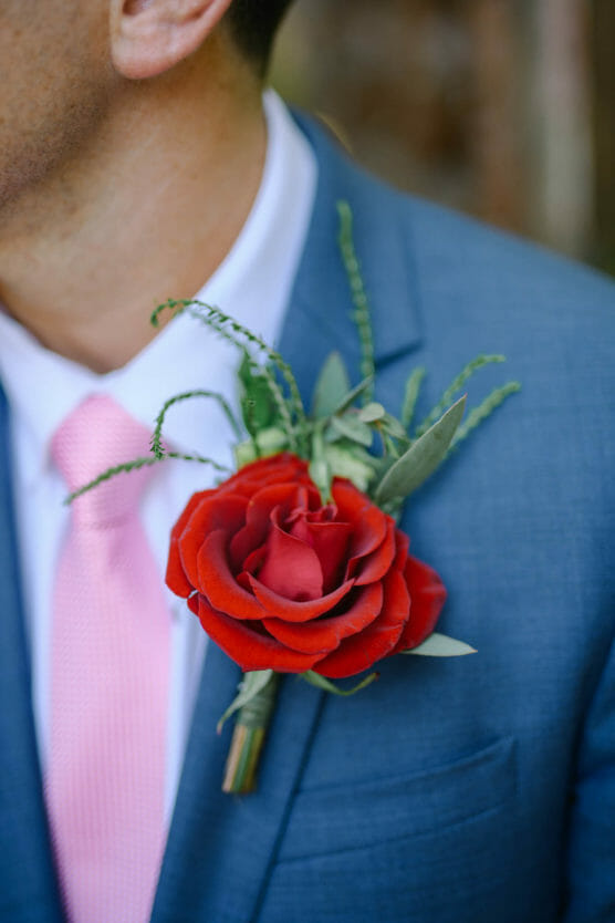 A close-up of a man wearing a blue suit and pink tie, with a red rose boutonniere and greenery pinned to his lapel. The mans face is partially out of frame. by Justin Salem Meyer