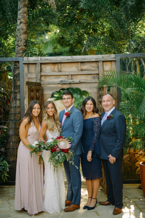 A bride and groom stand with three family members in formal attire, posing outdoors in front of a wooden gate surrounded by lush greenery. The bride holds a bouquet of flowers, and everyone is smiling. by Justin Salem Meyer