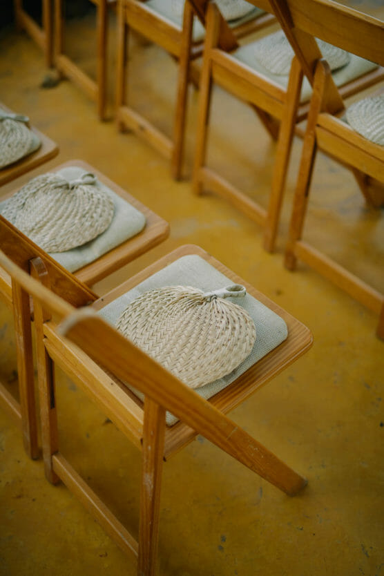 Wooden chairs arranged in rows, each with a woven hand fan placed on a cushion atop the seat, set on a yellowish floor. by Justin Salem Meyer