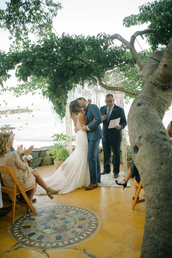 A bride and groom kiss under a tree during an outdoor wedding ceremony by the sea, with guests seated and an officiant standing behind them. The setting features a yellow floor and decorative mosaic tile. by Justin Salem Meyer