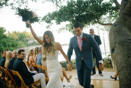 A bride and groom walk hand-in-hand down the aisle outdoors, smiling joyfully. The bride raises her bouquet in celebration as seated guests watch and smile. Trees and greenery surround the scene. by Justin Salem Meyer