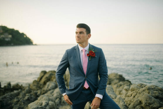 A man in a blue suit with a pink tie and a red rose boutonniere stands on rocky terrain by the sea, gazing to the side with a calm expression. The ocean and distant shoreline are visible in the background. by Justin Salem Meyer