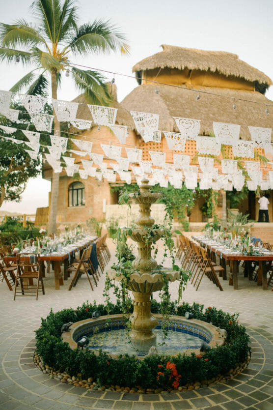 A decorative stone fountain with greenery is centered in a courtyard, surrounded by tables set for an outdoor event. Paper banners hang overhead, and a thatched-roof building and palm tree are in the background. by Justin Salem Meyer