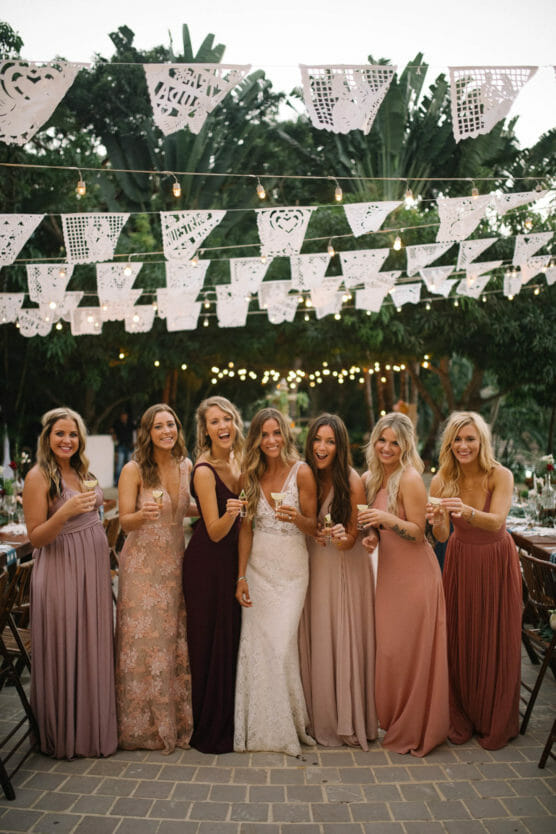 A bride in a white gown stands smiling with six bridesmaids in blush and mauve dresses, all holding drinks. They pose outdoors under hanging papel picado banners and string lights, with lush greenery in the background. by Justin Salem Meyer