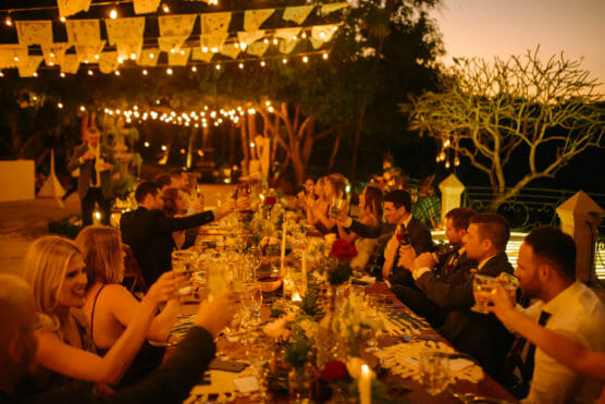 A group of people dressed formally raise their glasses in a toast at a long, outdoor dinner table, decorated with lights, lanterns, and greenery under string lights in the evening. by Justin Salem Meyer